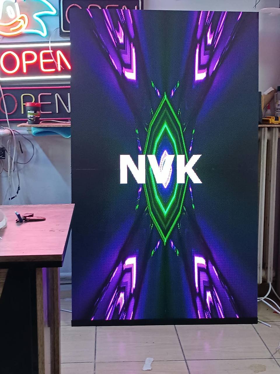 Atölyede NVK yazılı görüntü gösteren dikey LED ekran; arkada neon örnekleri.